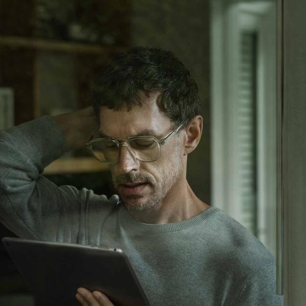 Man with glasses and a beard reading a tablet indoors, wearing a gray sweater and touching the back of his head.