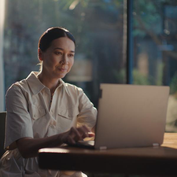 Person in a light-colored shirt working on a laptop at a wooden table, with a window showing greenery in the background.