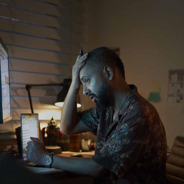 Man looking frustrated, sitting at a desk with a computer and notepad, resting his head on his hand in a dimly lit room.