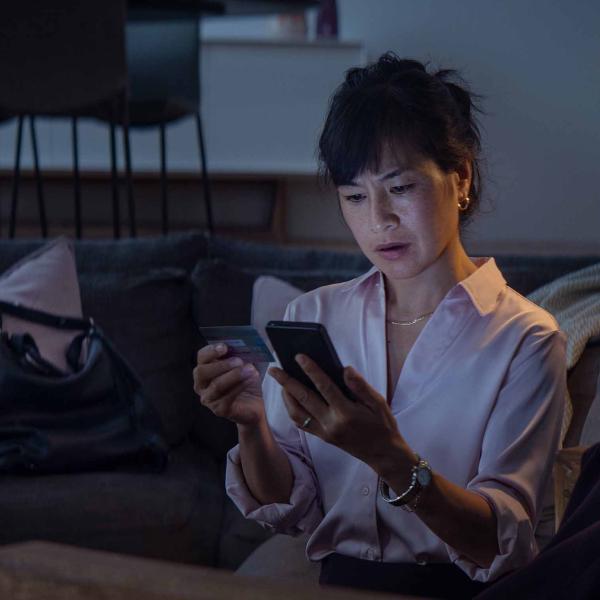 A woman sitting on a couch looks at her smartphone with concern, holding a credit card in her other hand.