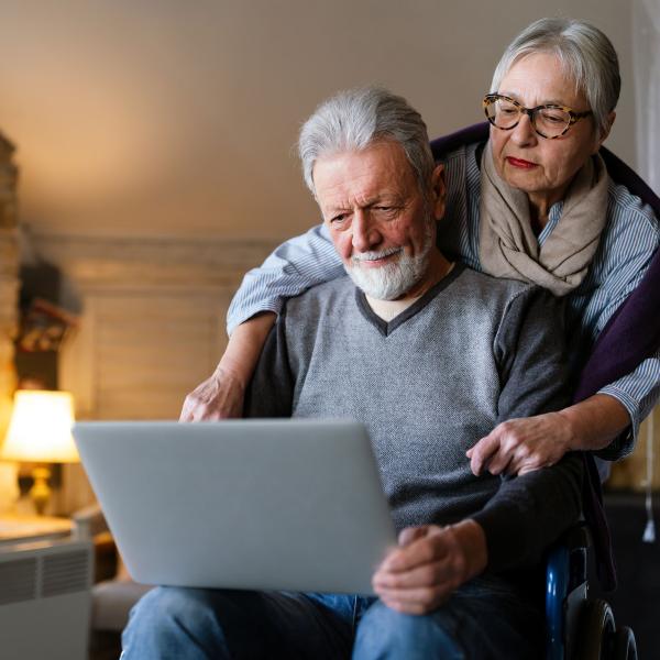 A man in a wheelchair using a laptop with an older women helping him update passphrases at home.