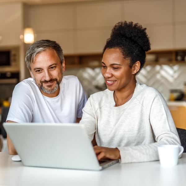 Two people sitting in a kitchen, looking at a laptop on the counter together.